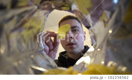 Young man eats potato chips while sitting at home. View of the man from the bag of chips. Young man eats potato chips while sitting at home. View of the man from the bag of chips. 102256562
