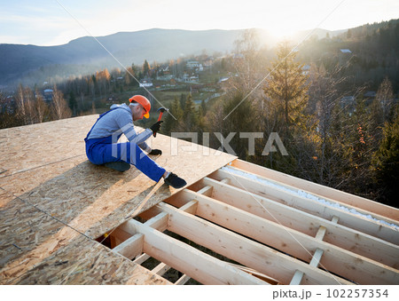 Carpenter hammering nail into OSB panel on the roof top of future cottage at sunset. Man worker building wooden frame house. Carpentry and construction concept. 102257354