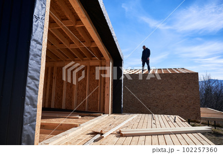 Male developer building wooden frame house. Bald man standing on construction site, inspecting quality of work on sunny day with blue sky on background. 102257360