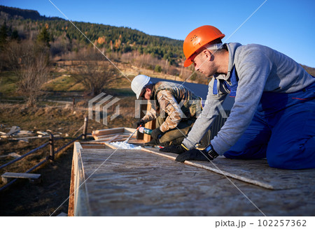 Male builder doing thermal insulation on roof of wooden frame house. Man worker spraying polyurethane foam on rooftop of future cottage. Construction and insulation concept. 102257362