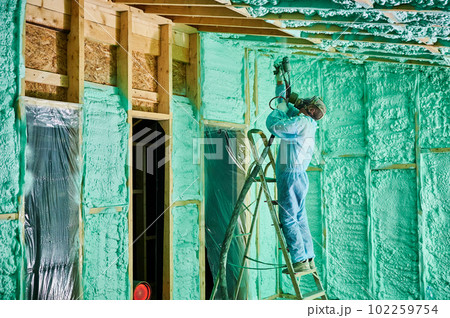 Male builder insulating wooden frame house. Man worker spraying polyurethane foam inside of future cottage, standing on ladder, using plural component gun. Construction and insulation concept. 102259754