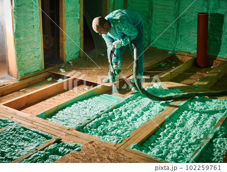 Male builder insulating wooden frame house. Man worker spraying polyurethane foam on floor inside of future cottage, using plural component gun. Construction and insulation concept. 102259761