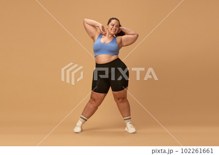 Smiling, positive, young overweight woman posing in comfortable sportswear against brown studio background. Caring about body Smiling, positive, young overweight woman posing in comfortable sportswear against brown studio background. Caring about body 102261661