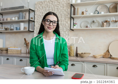 A young woman sits at home in the kitchen and writes with a pen in a notebook, keeps a personal diary, writes down dreams, makes a plan, works on self-developmente. He looks at the camera with a smile 102264361