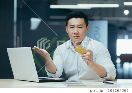 Worried young man sitting at desk in office and holding phone and blocked credit card, financial problems, unsuccessful shopping, bankruptcy. 102264362