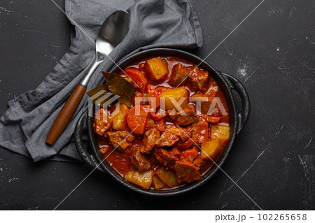 Beef meat stew with potatoes, carrot and delicious gravy in black casserole pot with bay leaves with spoon on black dark rustic concrete background from above . Beef meat stew with potatoes, carrot and delicious gravy in black casserole pot with bay leaves with spoon on black dark rustic concrete background from above . 102265658
