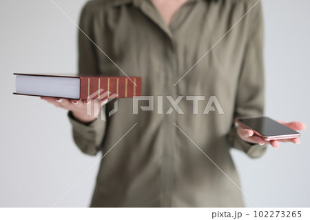 Confused woman holds book and smartphone making choice 102273265