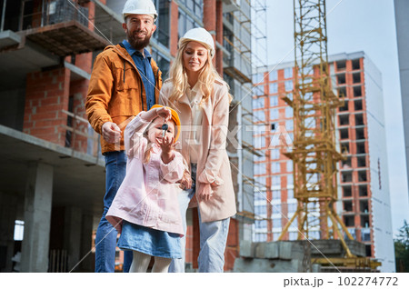 Daughter holding apartment keys and smiling while standing next to parents outside building under construction. Happy family homeowners posing on the street at construction site. 102274772