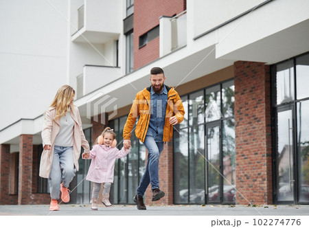 Full length of man and woman holding hands of little girl while walking down the street in new urban district. Happy loving family with child enjoying stroll outdoors near residential building. Full length of man and woman holding hands of little girl while walking down the street in new urban district. Happy loving family with child enjoying stroll outdoors near residential building. 102274776