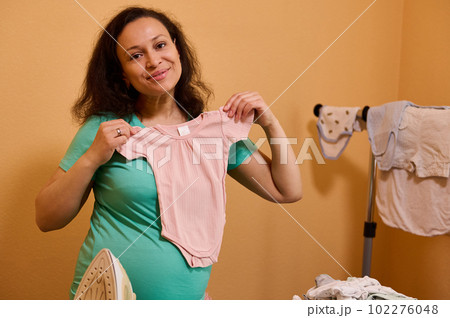 Happy expectant mother, pregnant woman smiling looking at camera, holding laundered clean ironed pink bodysuit for her newborn baby, standing by ironing board in dressing room. Household. Housekeeping 102276048