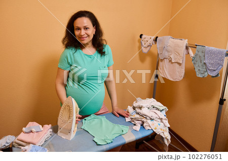 Beautiful middle aged multi ethnic happy pregnant woman with big belly, standing by ironing board with a stack of crumpled laundered baby clothes for her future newborn baby, smiling looking at camera Beautiful middle aged multi ethnic happy pregnant woman with big belly, standing by ironing board with a stack of crumpled laundered baby clothes for her future newborn baby, smiling looking at camera 102276051
