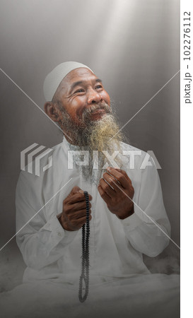 Muslim man with a beard wearing a white cap praying with prayer beads on his hands 102276112