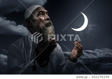 Muslim man with a beard wearing a white cap praying with prayer beads on his hands 102276149