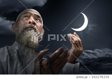 Muslim man with a beard wearing a white cap praying with prayer beads on his hands 102276150