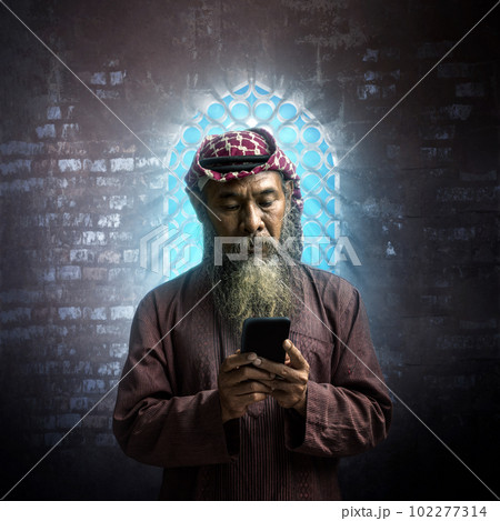 Muslim man with a beard wearing keffiyeh with agal using a mobile phone 102277314