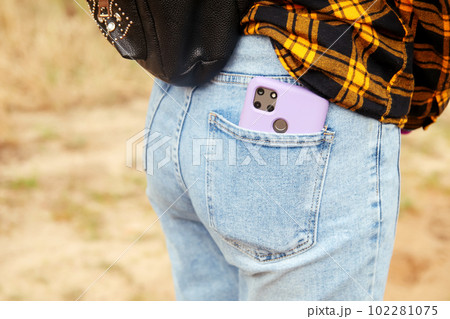 Woman puts mobile phone in jeans pocket on yellow nature background. Blue denim. Smart phone in the purple case. Out of focus 102281075