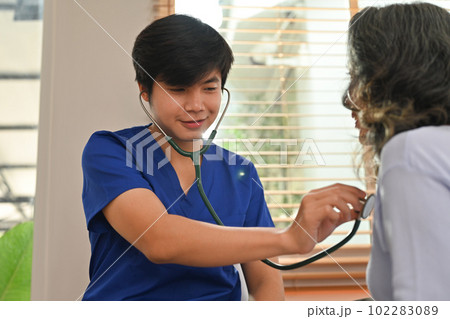 Male healthcare worker holding stethoscope checking heart and lungs of senior woman during home care visit Male healthcare worker holding stethoscope checking heart and lungs of senior woman during home care visit 102283089