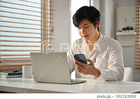 Handsome start up businessman holding smartphone and typing business email on laptop computer Handsome start up businessman holding smartphone and typing business email on laptop computer 102283092