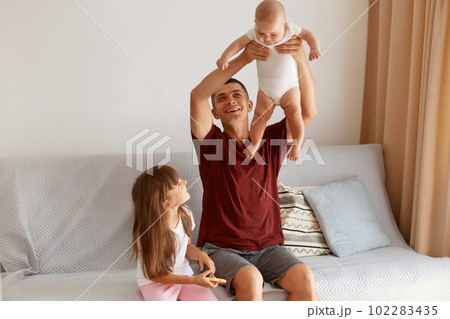 Indoor shot of brunette young adult Caucasian man wearing casual style burgundy t shirt posing in living room at home, sitting on sofa with his two kids, throwing up infant baby. Indoor shot of brunette young adult Caucasian man wearing casual style burgundy t shirt posing in living room at home, sitting on sofa with his two kids, throwing up infant baby. 102283435