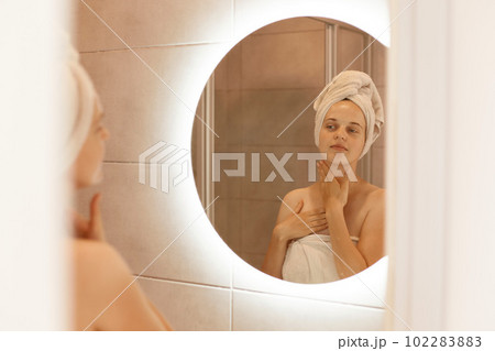 Indoor shot of beautiful woman looking on reflection in the mirror after shower, touching her neck, doing morning procedures, enjoying her fresh look. Indoor shot of beautiful woman looking on reflection in the mirror after shower, touching her neck, doing morning procedures, enjoying her fresh look. 102283883