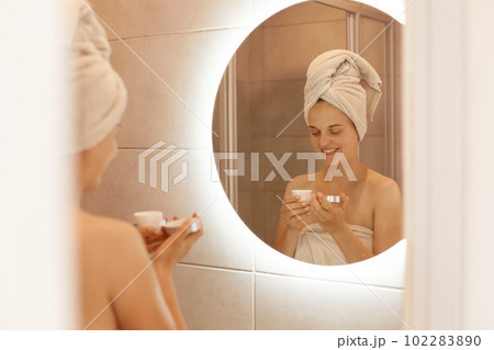Indoor shot of smiling woman moisturizes skin after the shower, holding cream in hands, expressing happy emotions, being wrapped in towel, doing spa procedures at home. 102283890