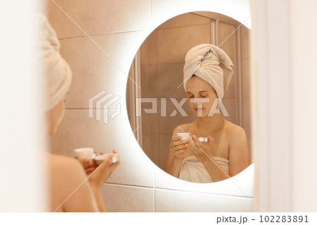 Portrait of young adult woman standing in bathroom in front of mirror with cream in hands, doing beauty procedures at home after taking shower, being wrapped in white towel. 102283891