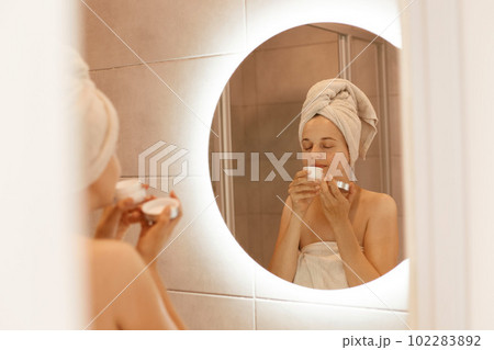 Young adult beautiful woman enjoying pleased cream odor, smelling cream before applying, standing with white towel on head in bathroom in front of mirror. 102283892