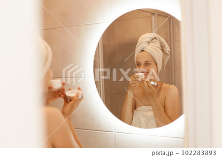 Charming female with perfect skin smelling cream and looking at her reflection in the bathroom mirror, standing with bare shoulders and white towel on her head. 102283893