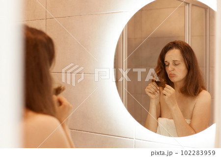 Indoor shot of young adult woman finding damaged hair, standing in front of a mirror in mirror, looking at dry hair ends, health care, treatment procedures. Indoor shot of young adult woman finding damaged hair, standing in front of a mirror in mirror, looking at dry hair ends, health care, treatment procedures. 102283959