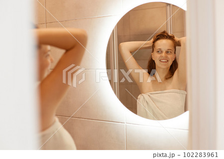 Smiling happy woman with perfect skin standing in front of mirror in bathroom with raised arms, morning routine, attractive female wrapped white towel. Smiling happy woman with perfect skin standing in front of mirror in bathroom with raised arms, morning routine, attractive female wrapped white towel. 102283961