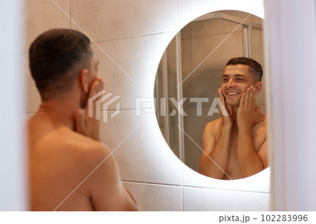 Happy positive brunette man standing in bathroom, looking at his reflection in the mirror, touching his cheeks, applying shaving agent on face, smiling. 102283996