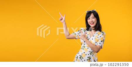 Portrait beautiful young asian woman wearing springtime dress and pointing to copy space for advertising isolated on yellow background. Summertime advertising banner concept. 102291350