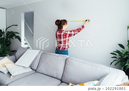 Young woman doing measuring with a measure tape on the wall. Girl wants to put a picture on the wall at home. Housekeeping work. Doing repair herself. DIY, gender equality in work concept. Young woman doing measuring with a measure tape on the wall. Girl wants to put a picture on the wall at home. Housekeeping work. Doing repair herself. DIY, gender equality in work concept. 102291801