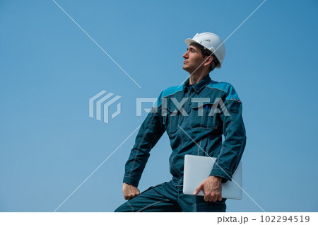 A male builder in a helmet stands against a blue sky with a laptop in his hands.  102294519