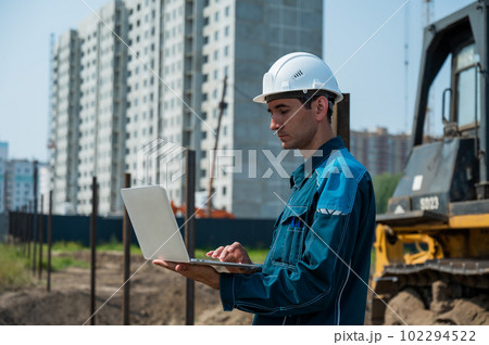 A male builder in a helmet stands against the background of a construction site with a laptop in his hands. A male builder in a helmet stands against the background of a construction site with a laptop in his hands. 102294522