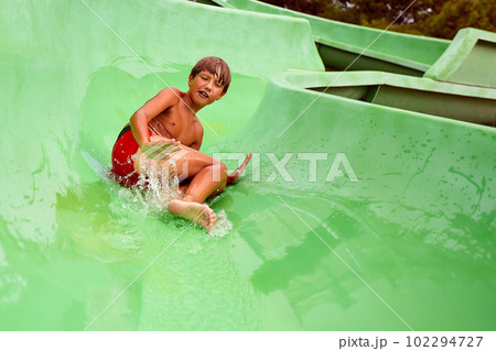 Kid descends from the water slide in the summer in the water park, happy child in the amusement park, summer family vacation. 102294727