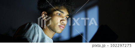 Evening routine. Portrait of focused young guy reading e book using tablet pc at night, sitting in dark room at home Evening routine. Portrait of focused young guy reading e book using tablet pc at night, sitting in dark room at home 102297275