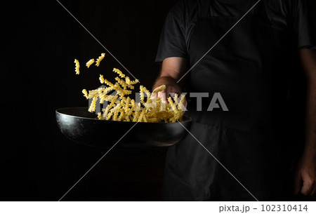 Professional chef prepares noodles in a hot frying pan with steam on a dark background. The concept of restaurant and hotel service Professional chef prepares noodles in a hot frying pan with steam on a dark background. The concept of restaurant and hotel service 102310414