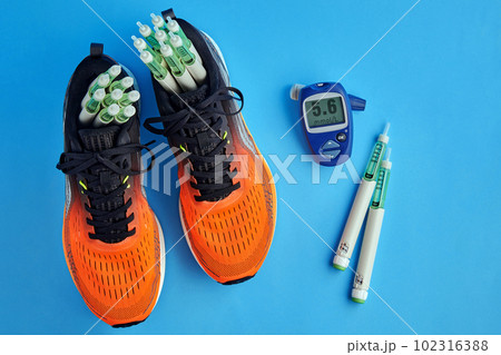 Orange running shoes with insulin syringe pens and a glucometer for measuring blood sugar surrounded by decorative red hearts on a blue background 102316388