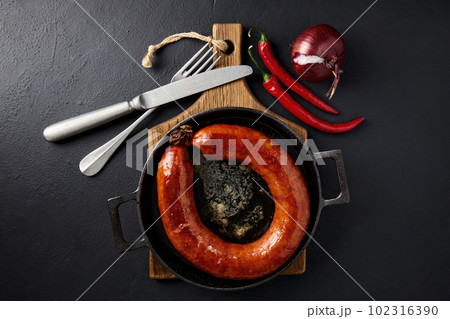 Fried sausage rolled into a ring on a cast iron pan with cutlery, a wooden cutting board, hot peppers and red onions 102316390