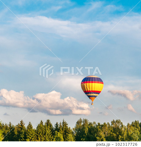 A bright hot air balloon flies in the blue sky above the treetops of the forest. Bright warm colors 102317726