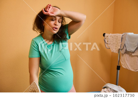 Portrait of an adult pregnant woman removing sweat from her forehead, feeling tired and overworked by household chore, ironing baby clothes, preparing for the birth of child. Pregnancy and Maternity Portrait of an adult pregnant woman removing sweat from her forehead, feeling tired and overworked by household chore, ironing baby clothes, preparing for the birth of child. Pregnancy and Maternity 102317730