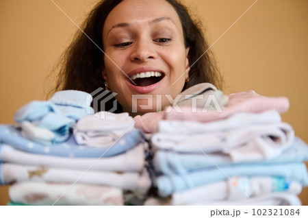Exhausted but happy woman, smiling with a beautiful toothy smile, holding clean ironed baby clothes, enjoying happy moments for her pregnancy and maternity leave. Preparing for the birth of her child Exhausted but happy woman, smiling with a beautiful toothy smile, holding clean ironed baby clothes, enjoying happy moments for her pregnancy and maternity leave. Preparing for the birth of her child 102321084