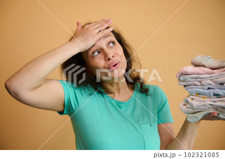 Tired, exhausted, overworked and multitasking pregnant woman, housewife, mother puts her hand on forehead, feels tiredness after household work, holds ironed laundered baby clothes on color background 102321085