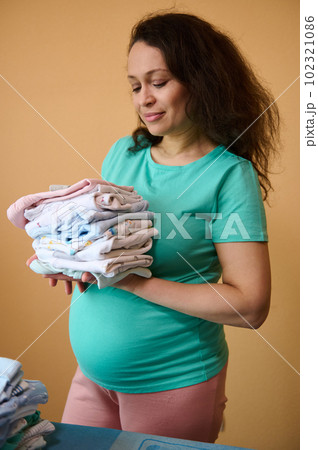 Vertical studio portrait of a confident adult multi ethnic pregnant woman, holding clean laundered ironed clothes for her future newborn baby, standing at ironing board over yellow wall background Vertical studio portrait of a confident adult multi ethnic pregnant woman, holding clean laundered ironed clothes for her future newborn baby, standing at ironing board over yellow wall background 102321086