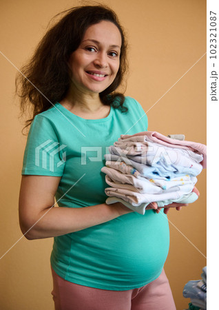 Cheerful expectant mother, pregnant woman smiling broadly looking at camera, holding a stack of clean, laundered and ironed newborn baby clothes on her hand, isolated over orange color background 102321087