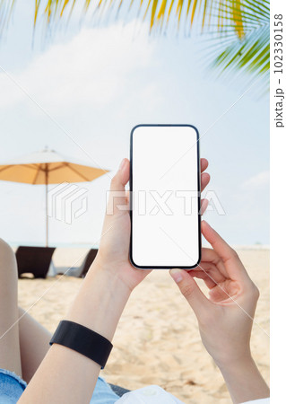 Woman sitting on deckchair on the beach with hand holding smartphone mockup of blank screen. 102330158
