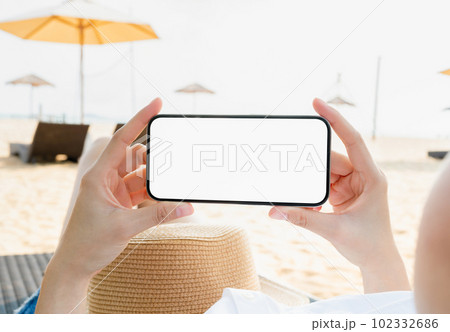 Woman sitting on deckchair on the beach with hand holding smartphone mockup of blank screen. 102332686