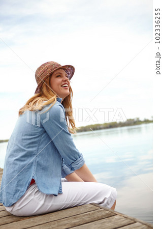 Relaxing by the waters edge. A happy young woman sitting on a jetty next to a lake. 102336255