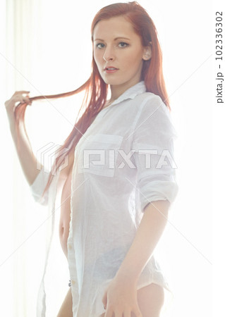 Shes a firery beauty. Portrait of an attractive redheaded woman wearing an unbuttoned shirt standing in a bedroom looking sexy while standing in her underwear. Young female posing semi nude. 102336302